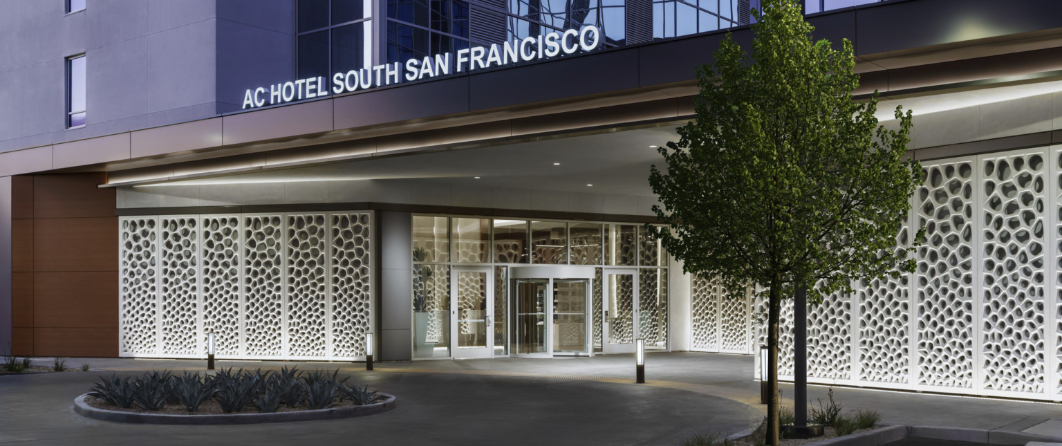 Exterior view of hotel entry with illuminated sign above. White organic sculpture lines walls under driveway shelter at entry