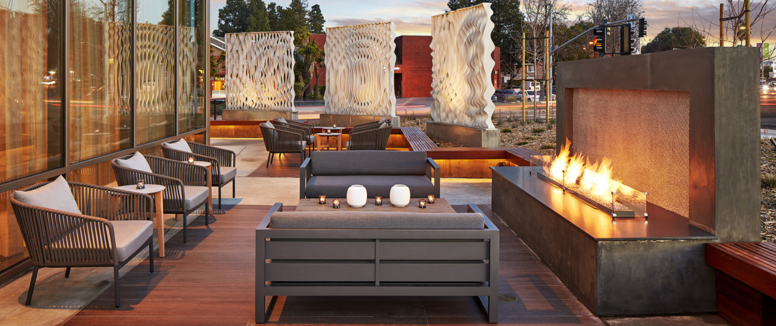 Outdoor seating area with couches and arm chairs in front of fireplace. Behind, more seating with 3 white organic sculptures