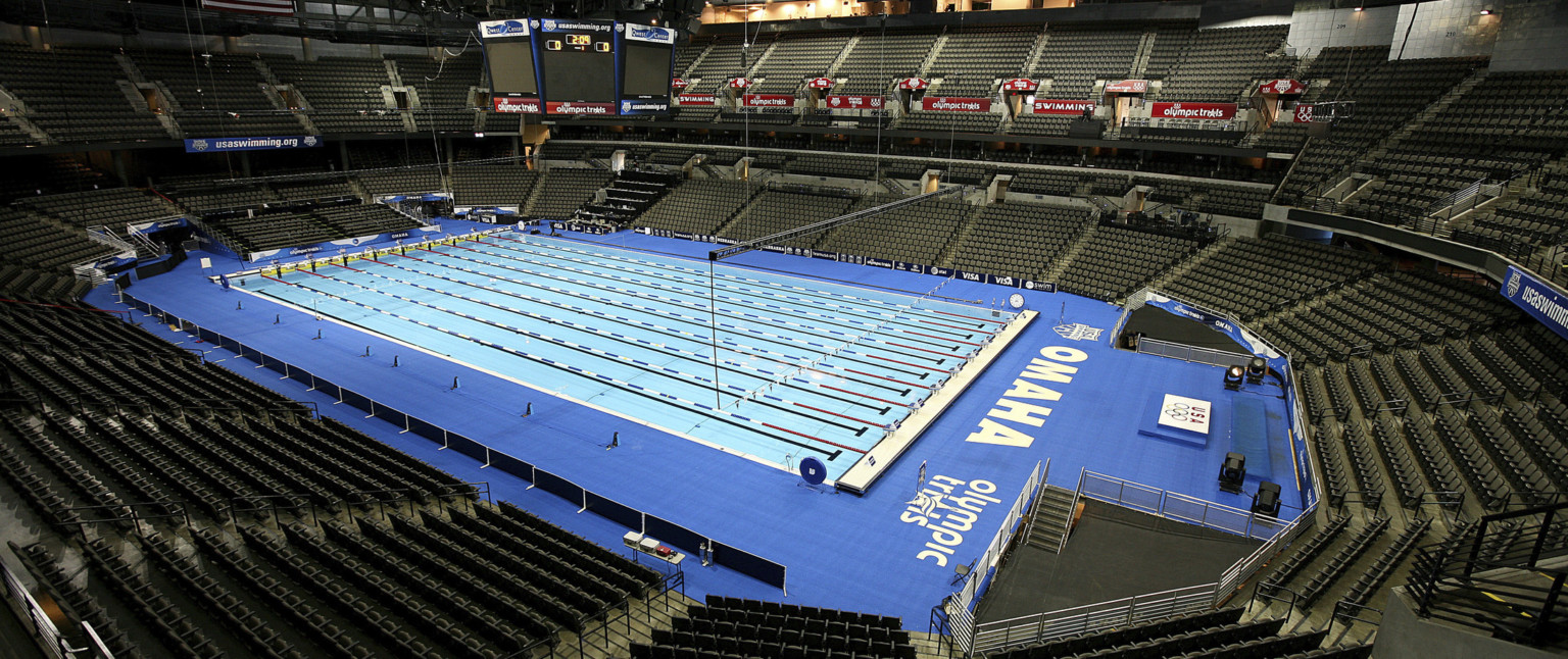 A temporary Olympic sized swimming pool set up within the empty arena. Jumbotron above and poolside have Olympic trials signage