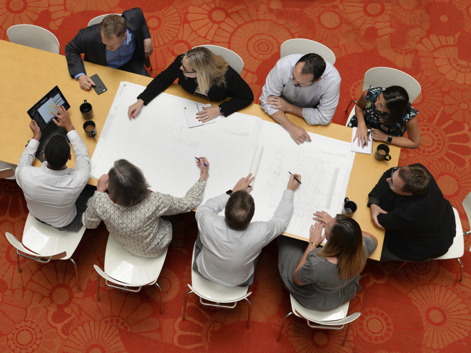 Aerial view of group working at wood conference table with grey chairs on red floral rug. White paper and tablet in use