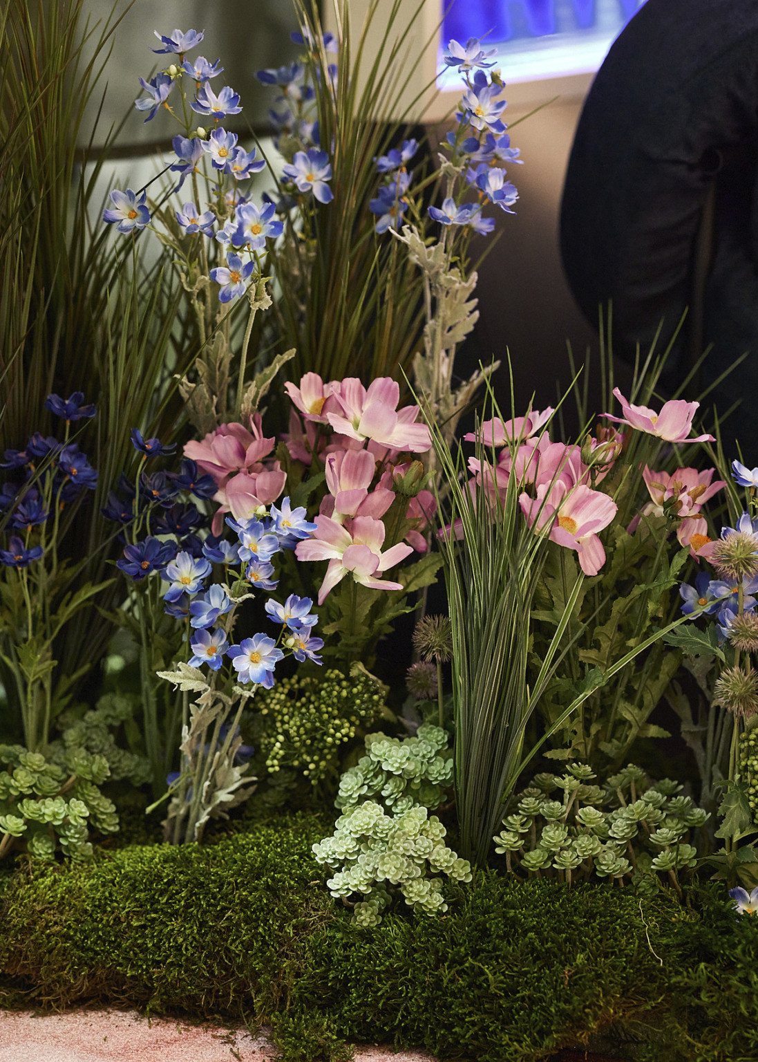 Close up of faux florals placed on ground. Fake flowers in blue, pink, and purple with synthetic moss and grass
