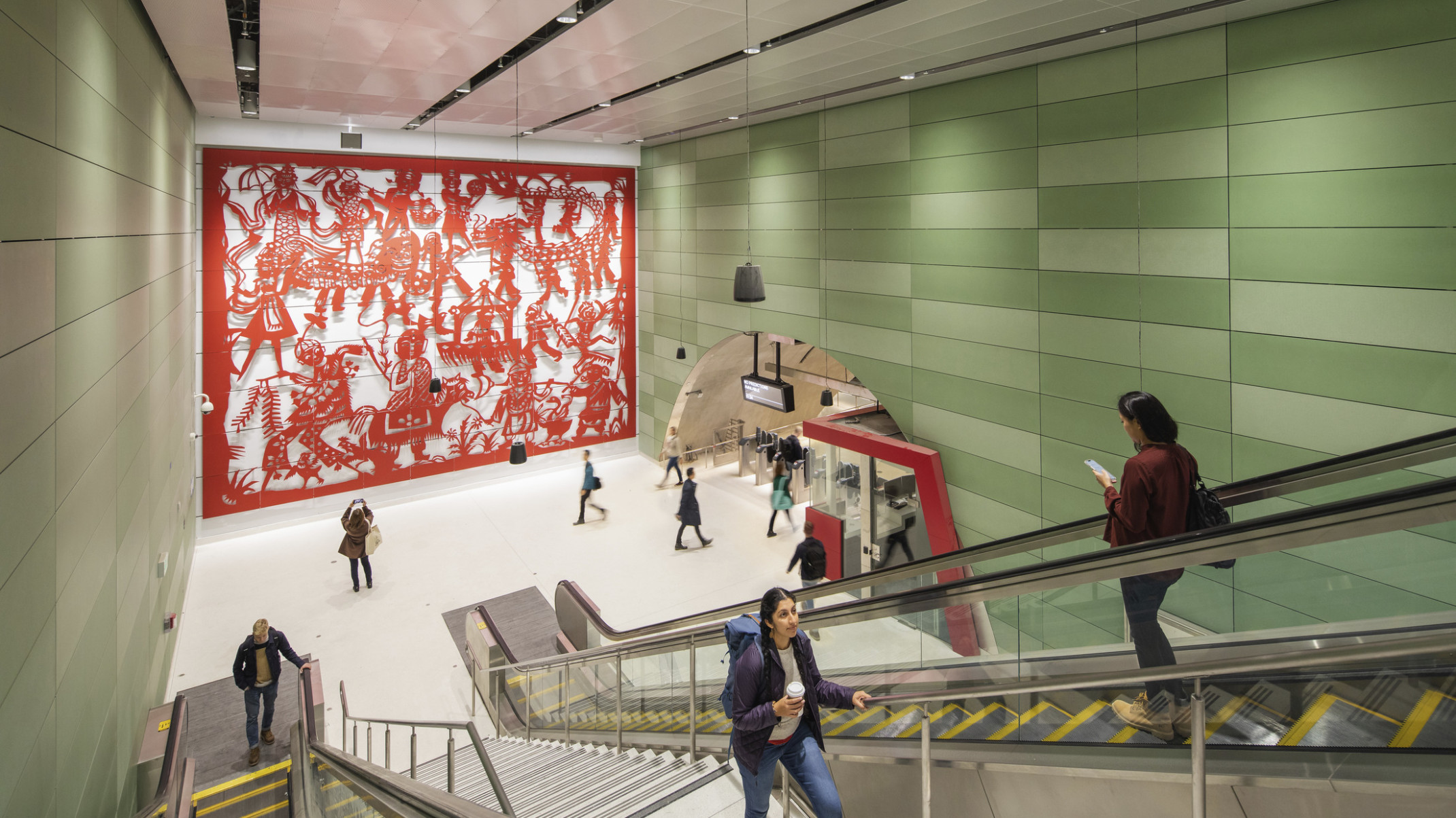 Concrete block walls with raised red sculptural mural on left. Right, arched hall with gates and ticket booth by escalators