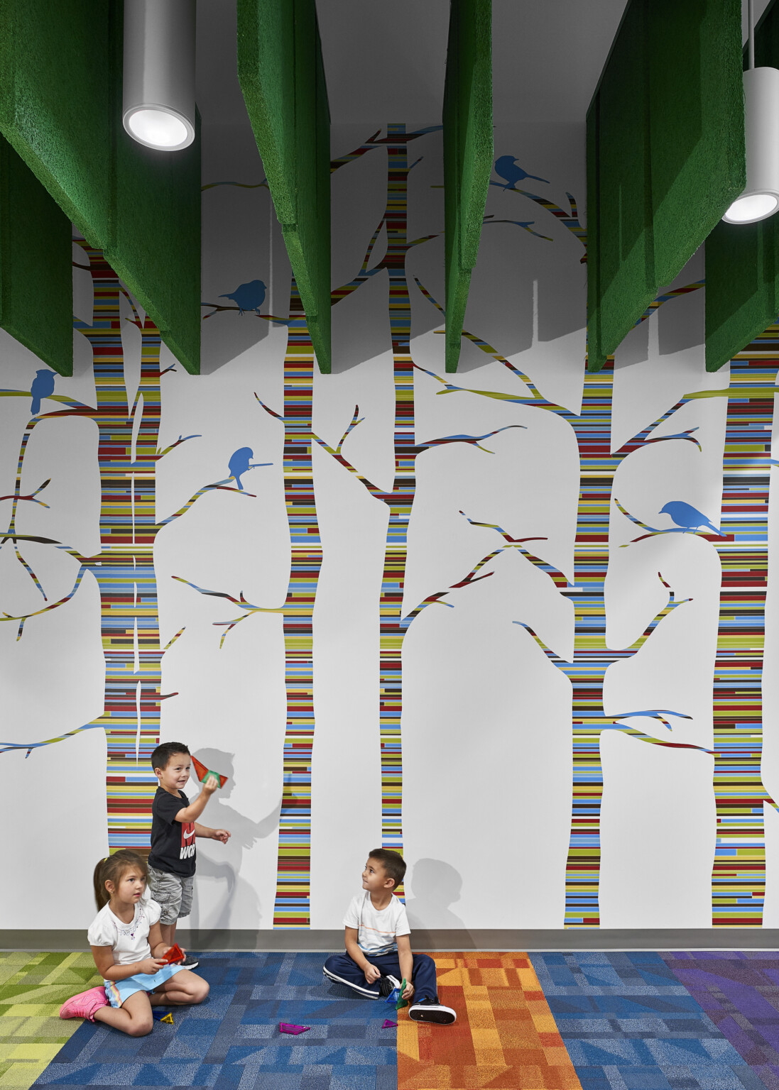 Students sit on multicolor carpet in front of striped tree mural with blue birds. Green acoustic panels hang above