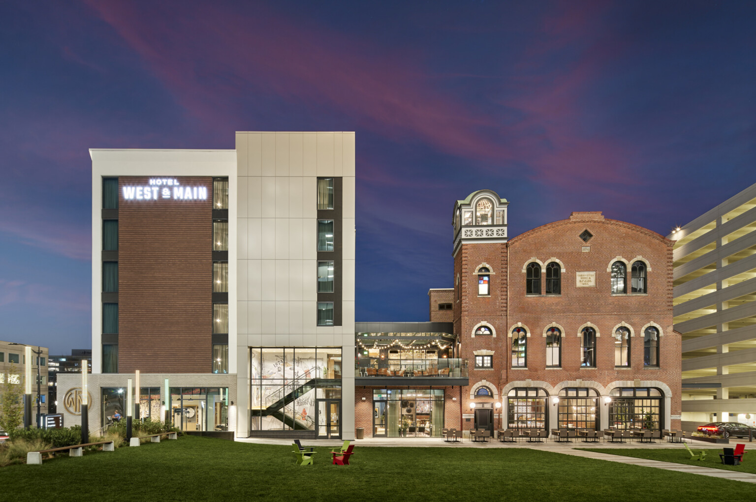 On the left, a new multi-story hotel wrapped in light siding with brick accents and on the right, the former brick fire station connects to the new hotel by an enclosed structure with a rooftop balcony overlooking a lawn