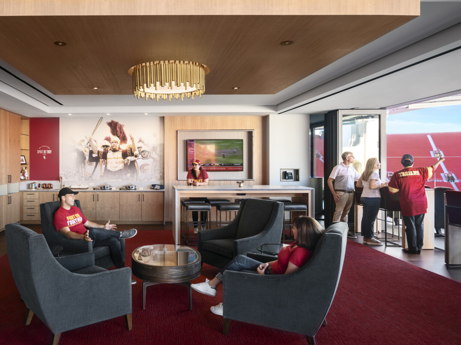 Private box in stadium with red carpet and gold chandelier. Flexible wall folds open facing field. Counter space and seating