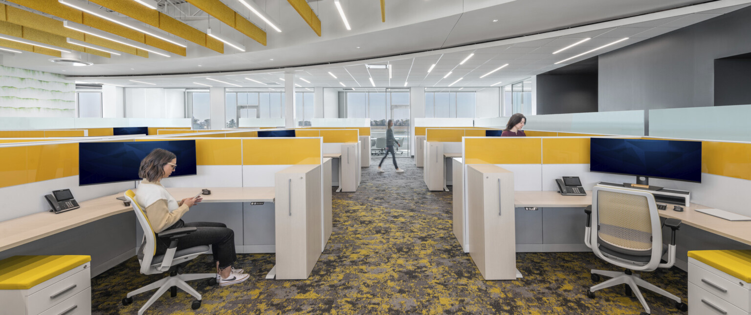 Open workspace with sit-stand desk and pops of yellow on the workstations, carpet, and ceiling baffles