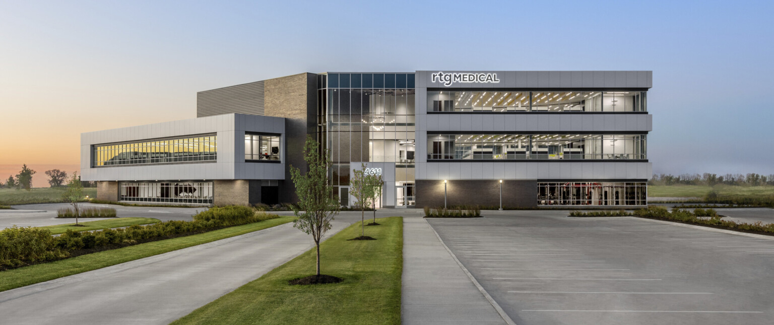 Front side of 3 story building illuminated in the early morning. Green space surrounding. Grey wrap facade accent, large windows Front side of 3 story building illuminated in the early morning. Green space surrounding. Grey wrap facade accent, large windows