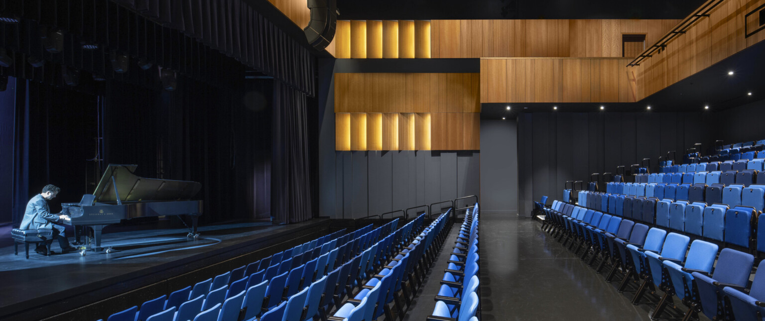 Theater with black walls, wood sections at top and balcony, facing a illuminated stage with pianist playing; blue seating