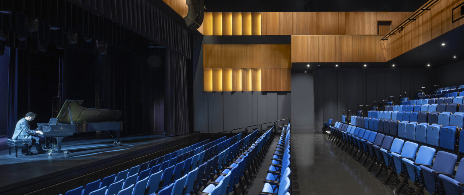 Theater with black walls, wood sections at top and balcony, facing a illuminated stage with pianist playing; blue seating Theater with black walls, wood sections at top and balcony, facing a illuminated stage with pianist playing; blue seating