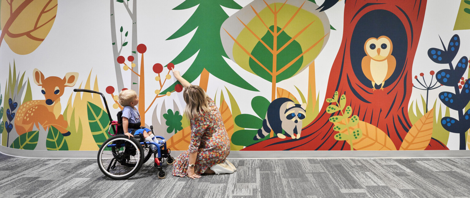 Child and educator interact with mural of cartoon forest and animals in carpeted school hallway. Mixed trees, deer, owl, racoon