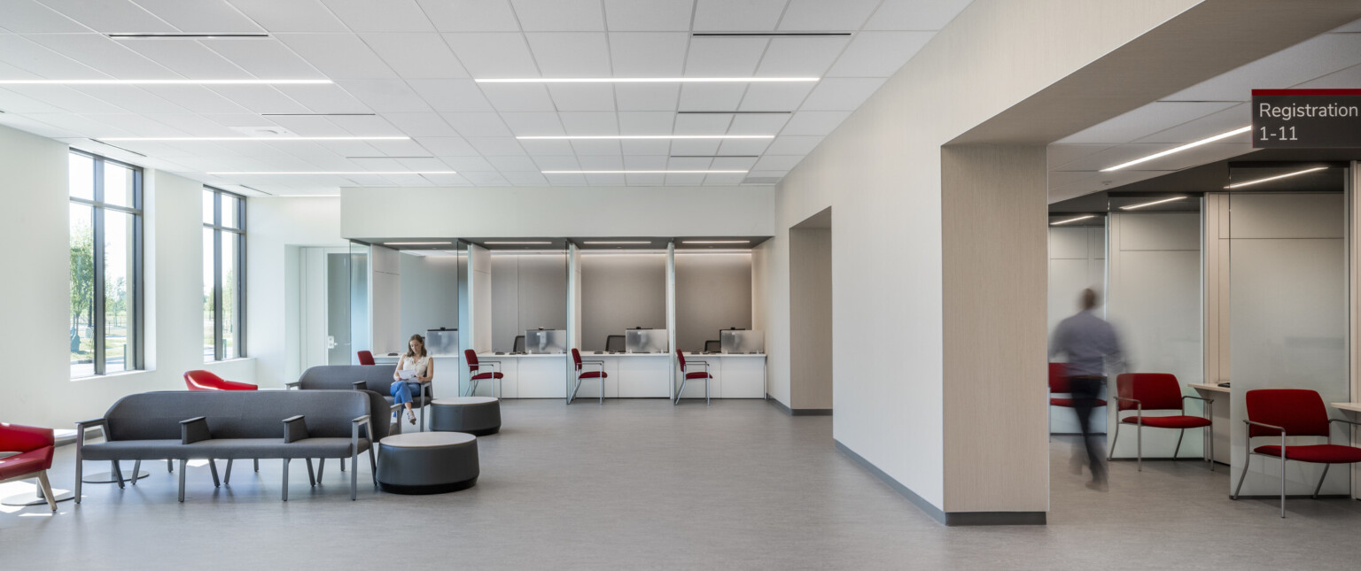 White waiting room with wall separating seating area with grey chairs from registration area with red chairs and glass partitions