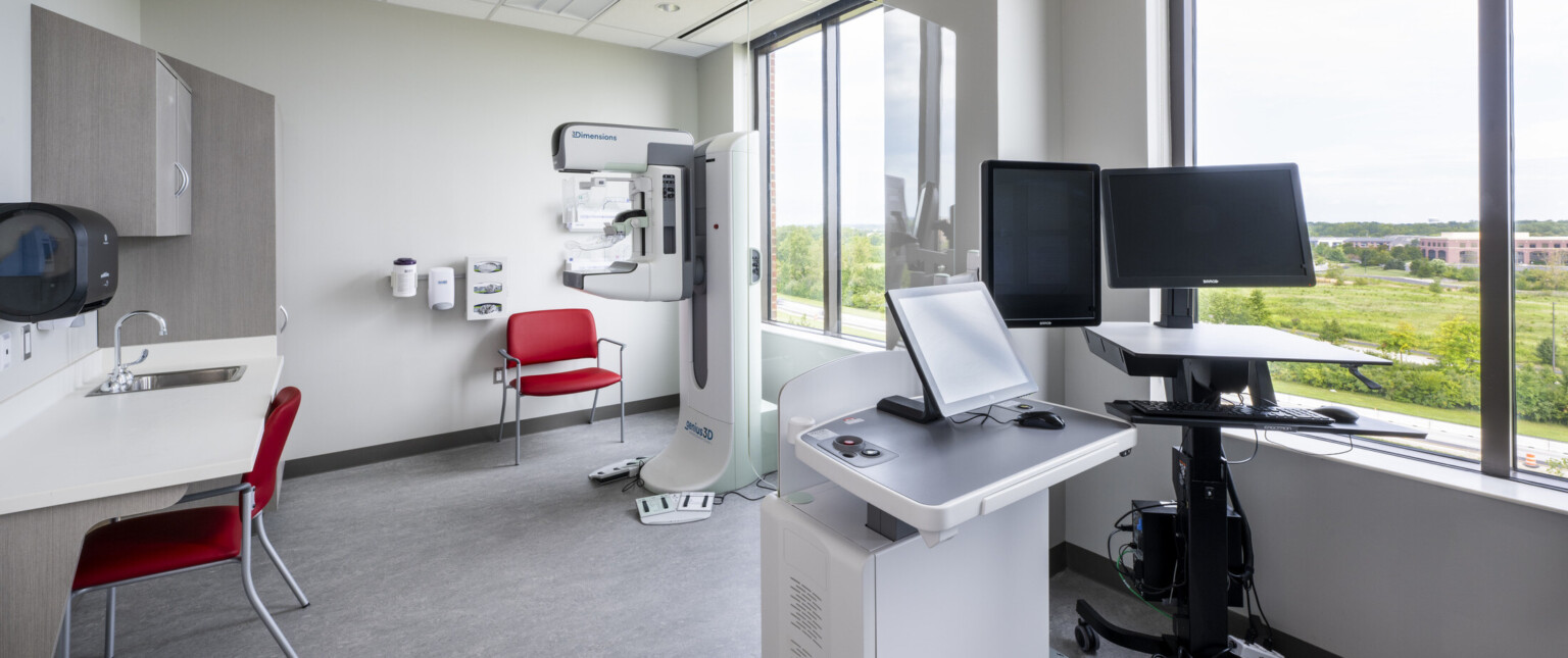 Patient care space with sink, portable standing desk with computer monitors, healthcare equipment. Large windows on right wall