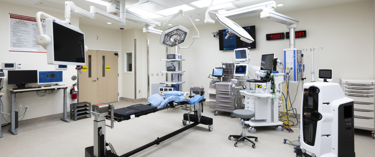 Operating room with medical equipment along outside edge of white room with two digital wall clocks. Equpment hangs above