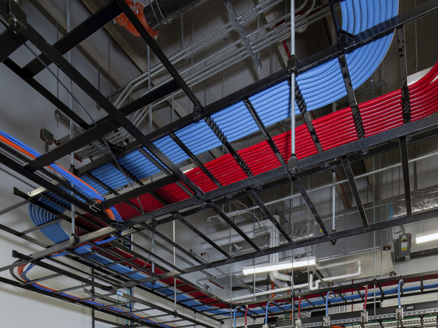 an open ceiling with black orthogonal framing system that contains lighting pendants, pipes, metallic and red and blue tubes