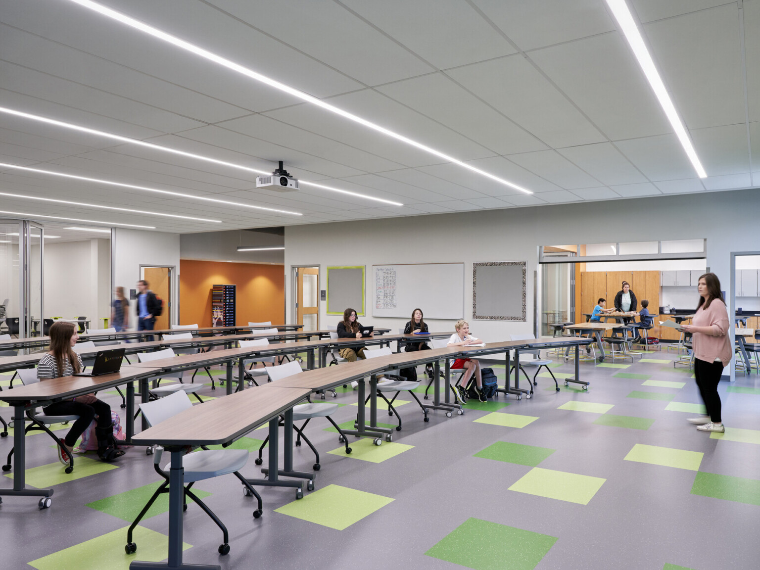 Classroom with green accent wall and floor tiles. Orange walls in hallway through flexible walls. Rolling chairs and long tables