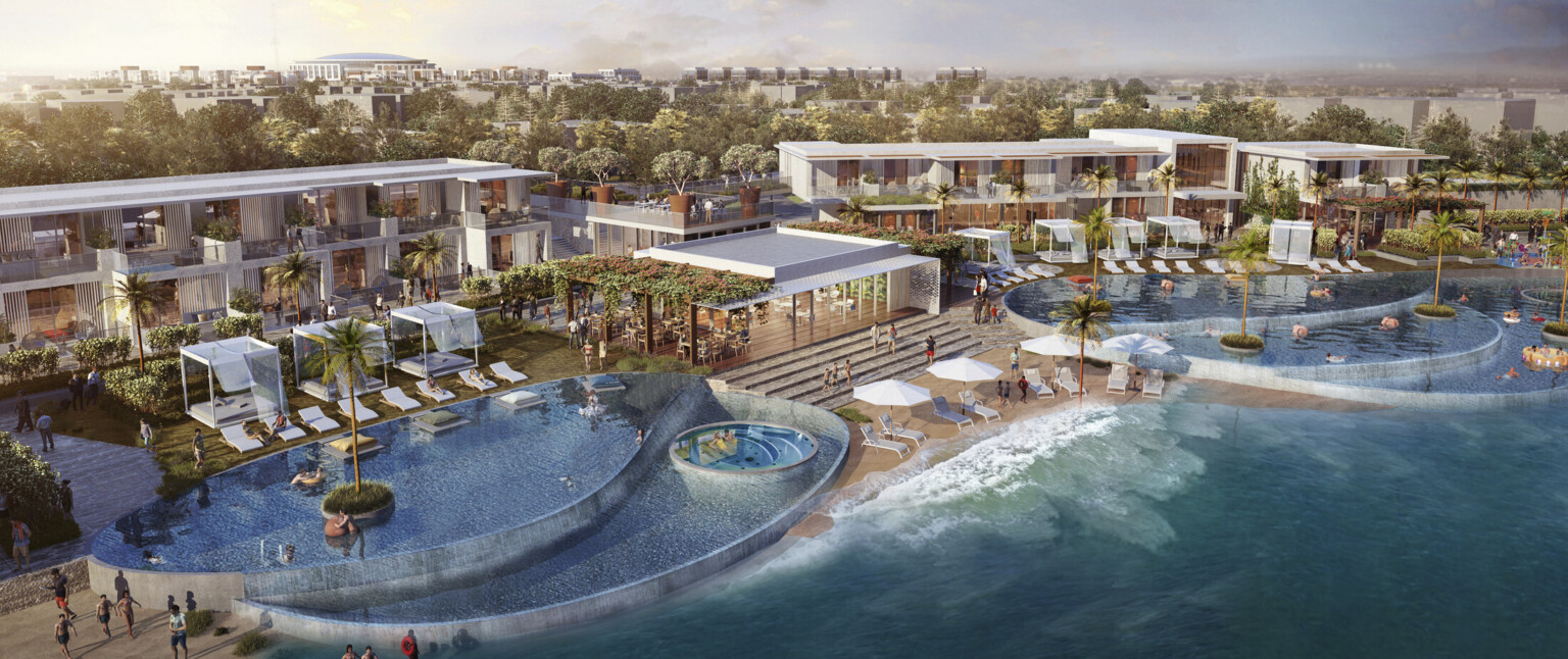 Multiple pools cascading to water on either side of small beach lounge space and large outdoor area. Buildings and trees behind