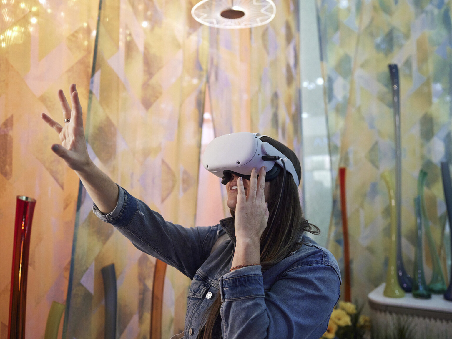 Woman using VR headset reaches out in room with geometric triangular patterned curtains in warm cool tone gradient. Organically shaped vases