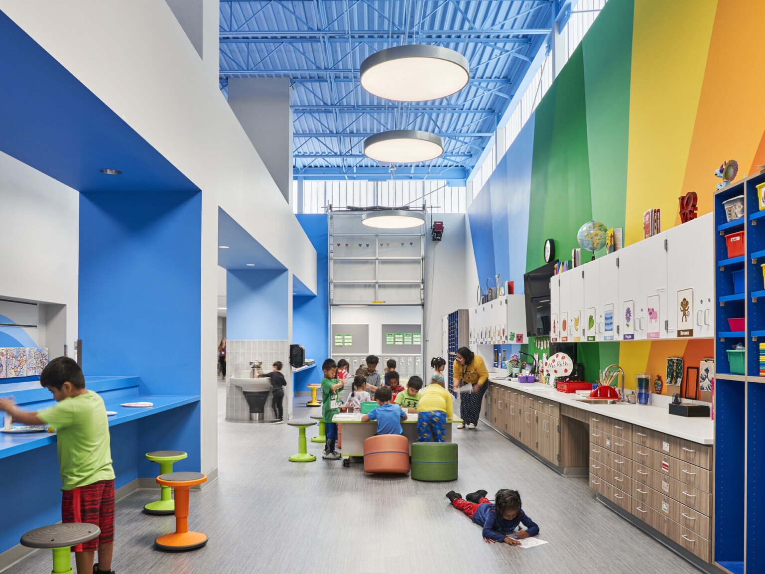 Classroom interior open-concept classroom with vaulted ceilings, custom lighting fixtures, natural daylight, brightly colored walls, children in various seating arrangements