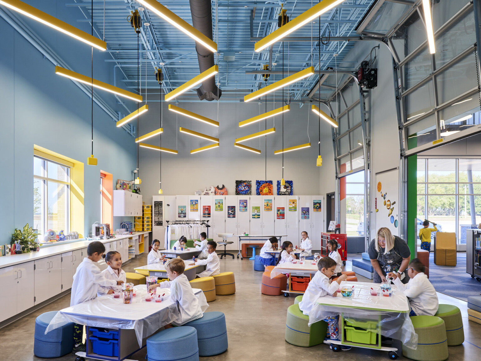 Classroom interior open-concept classroom with vaulted ceilings, custom lighting fixtures, brightly colored walls, natural daylight, children seated at worktables with colorful, ergonomic seating arrangements