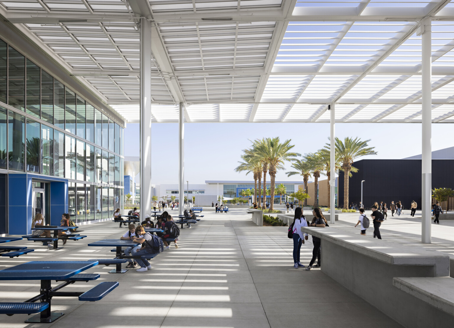 Outdoor seating area with white canopy overhead. Glass wall left with blue accent around doors. View of courtyard and palm trees