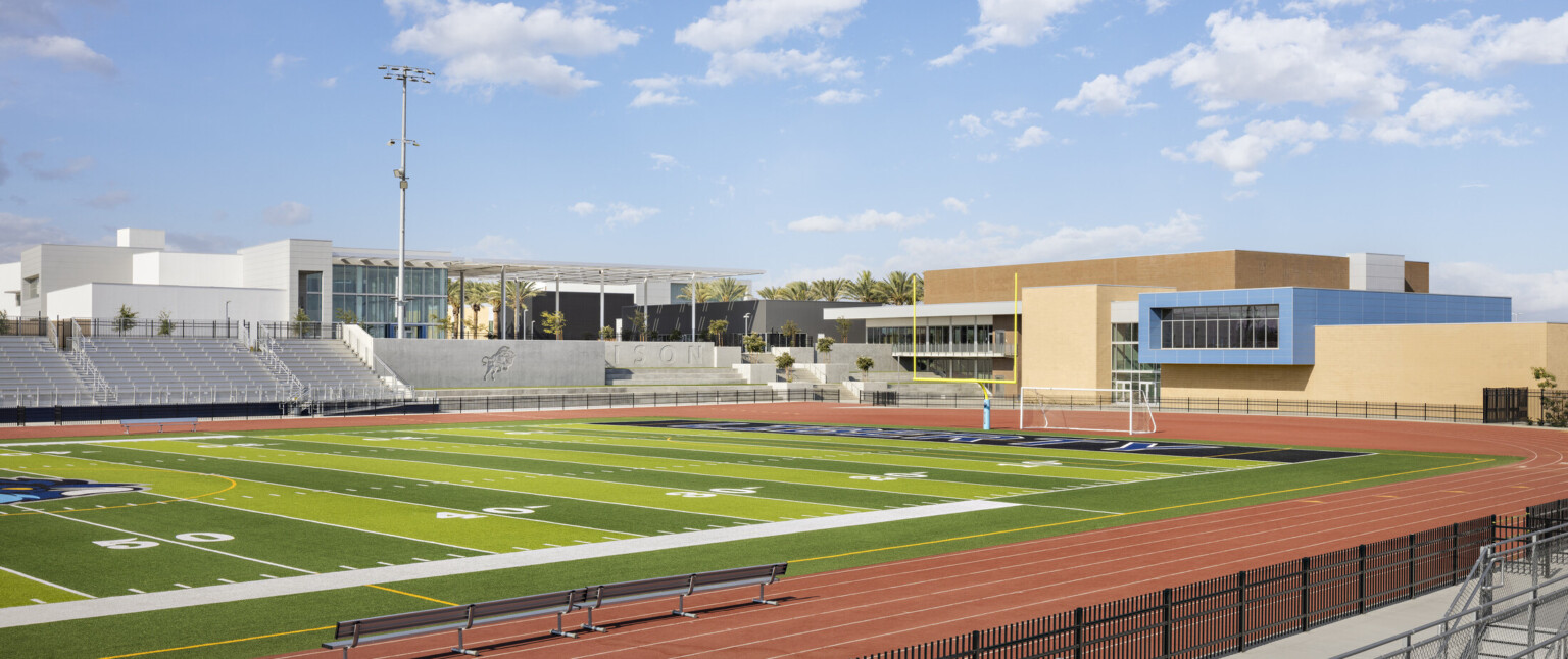 Football field surrounded by track with white bleachers, school beyond with blue accent hallway with floor to ceiling windows