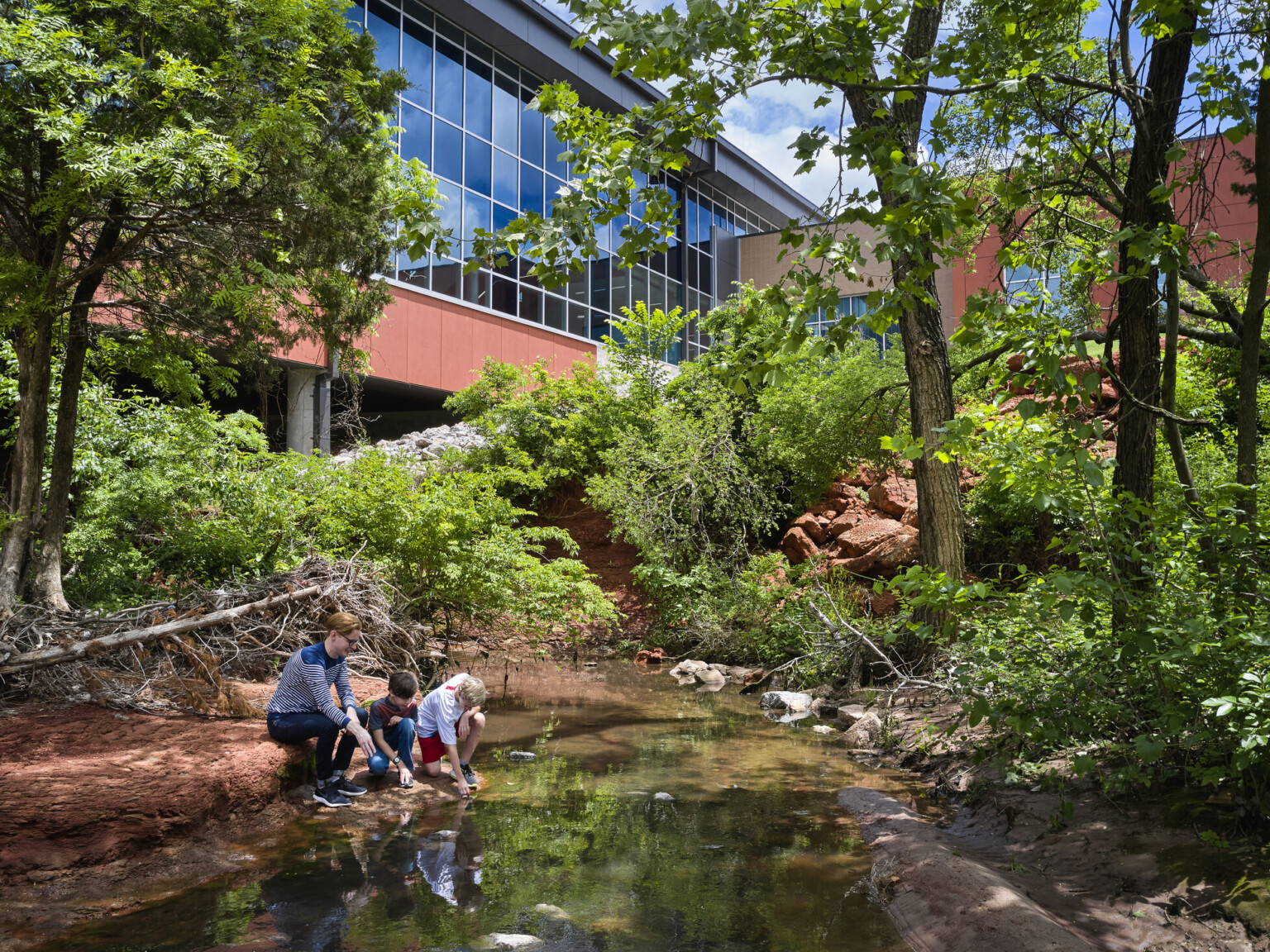Two young students participating in outdoor learning near modern structure with floor-to-ceiling windows, lush landscaping and a reflecting pond Two young students participating in outdoor learning near modern structure with floor-to-ceiling windows, lush landscaping and a reflecting pond
