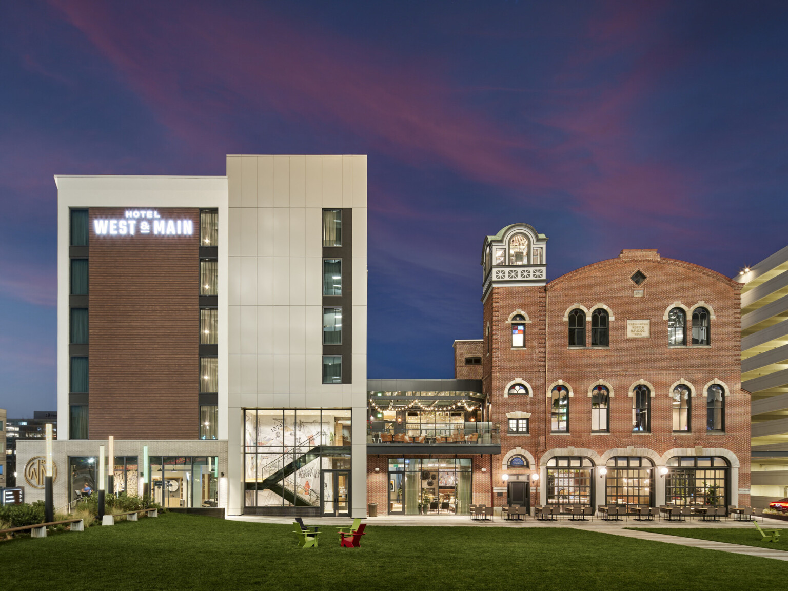 On the left, a new multi-story hotel wrapped in light siding with brick accents and on the right, the former brick fire station connects to the new hotel by an enclosed structure with a rooftop balcony overlooking a lawn
