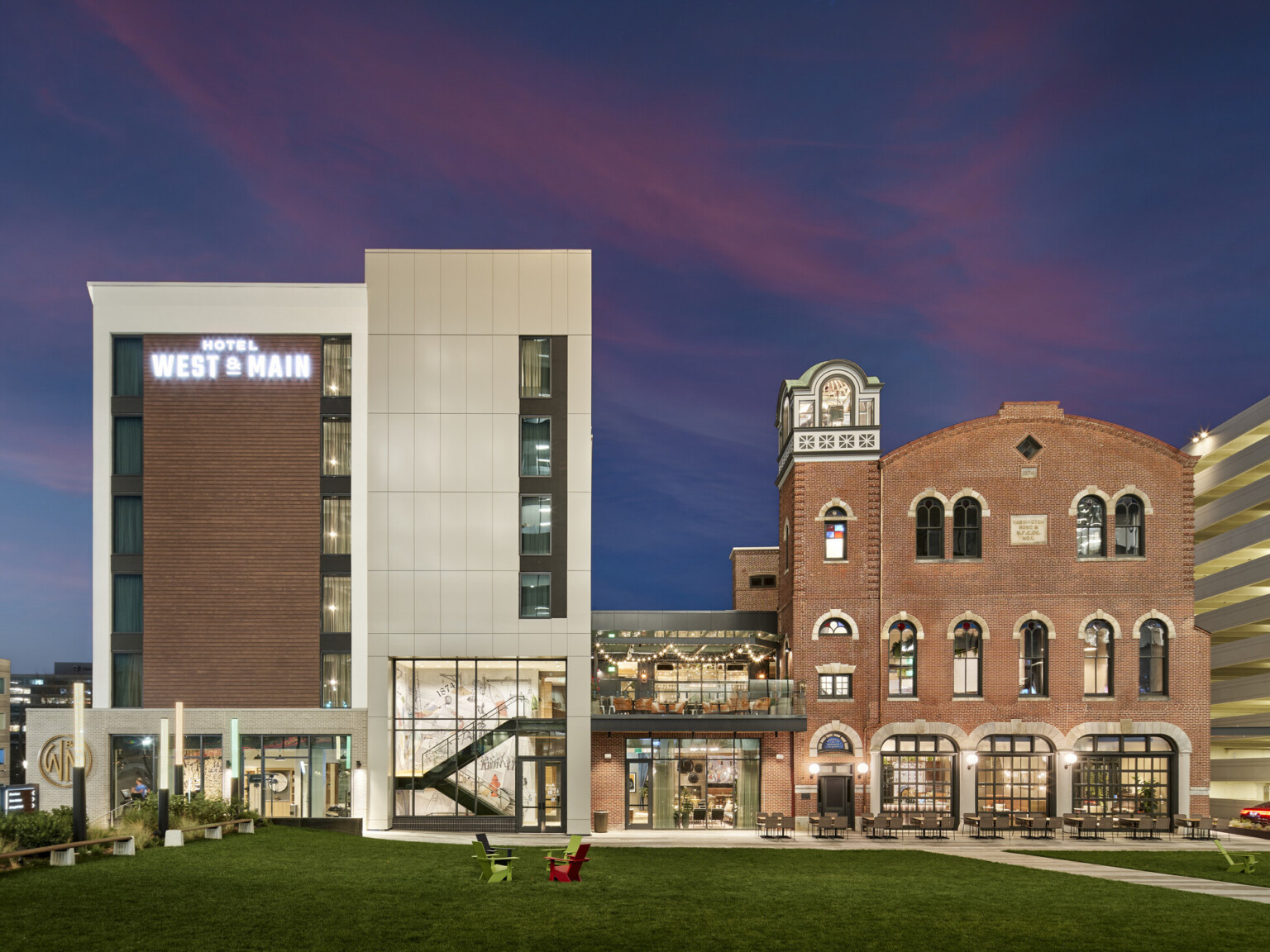 On the left, a new multi-story hotel wrapped in light siding with brick accents and on the right, the former brick fire station connects to the new hotel by an enclosed structure with a rooftop balcony overlooking a lawn On the left, a new multi-story hotel wrapped in light siding with brick accents and on the right, the former brick fire station connects to the new hotel by an enclosed structure with a rooftop balcony overlooking a lawn
