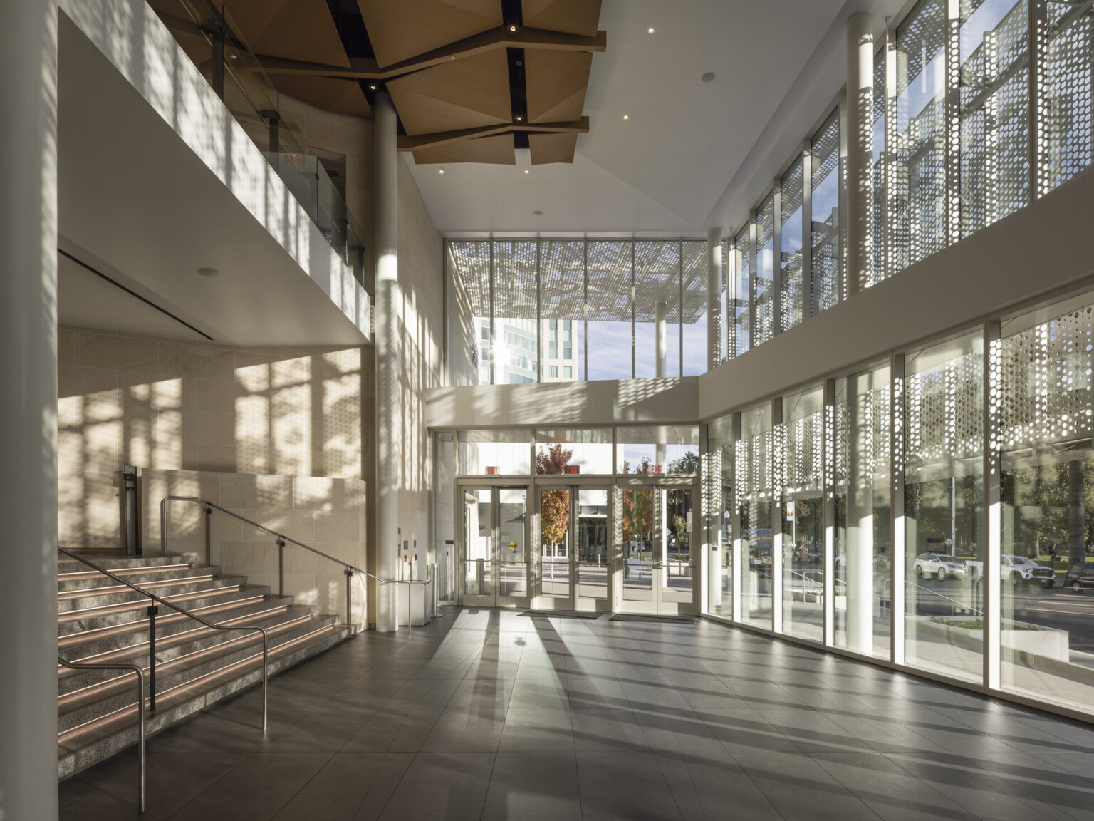 Two-story lobby space lit by floor to ceiling glass windows; wood sculpture covers ceiling; perforated light shines through the exterior lacy scrim