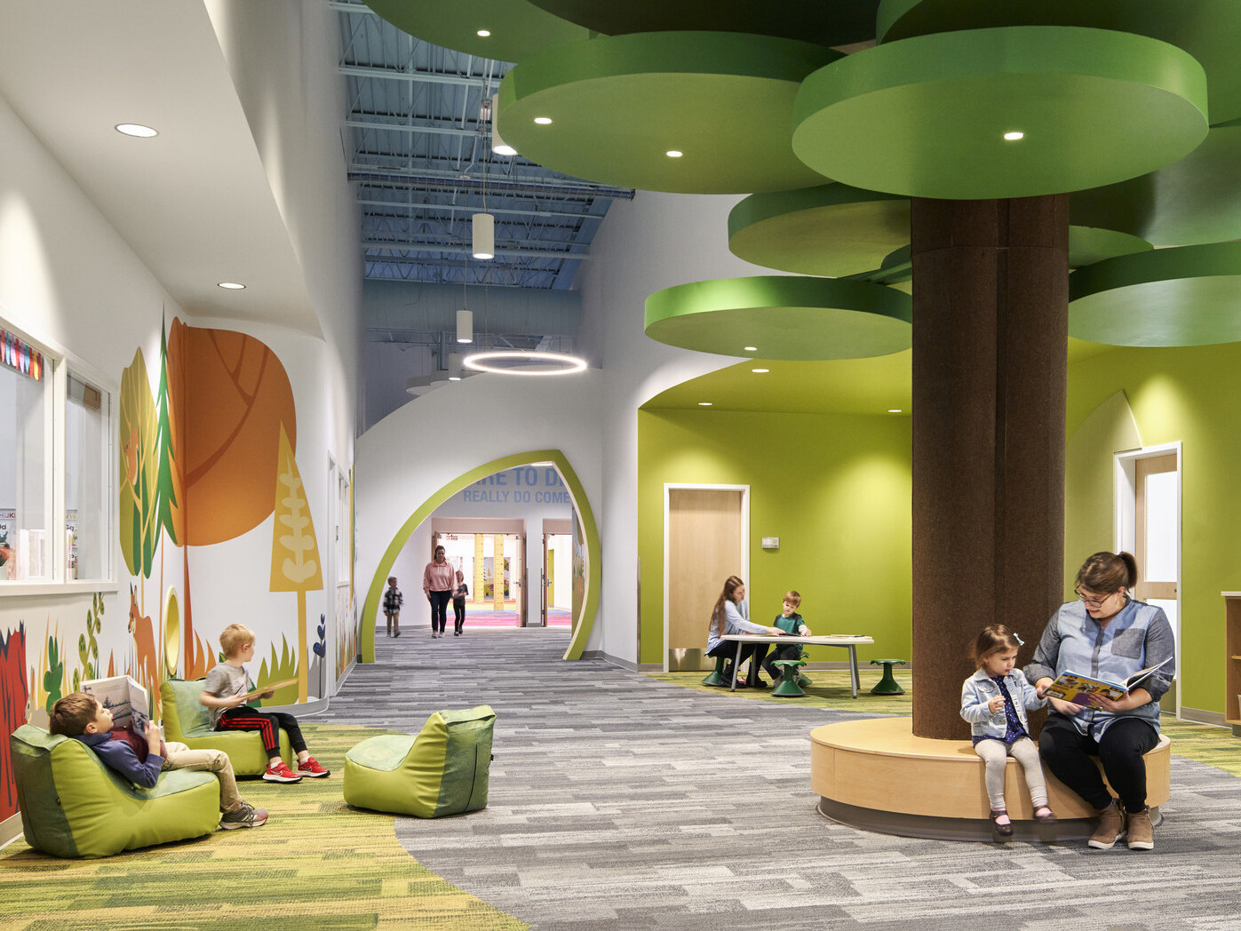 people reading on a brown circular bench with a brown column and green ceiling fixtures in hallway lined with forest graphics